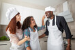 © sofiko14 - Multiethnic family in aprons and hats, baking in the kitchen. Cute curly-haired kid girl, touching her father's face with hands stained with flour