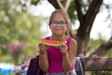 Girl eating a piece of watermelon on a bench in a sunny summer garden. She smiles and enjoys the juicy sweet watermelon