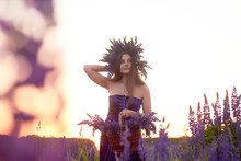 A Young Girl With A Floral Wreath On Her Head Stands At Sunset In A Field, Holding A Wicker Basket With A Large Luxurious Bouquet With Purple Lupins. Blooming Field In Summer.