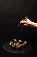 Wall Mural - Hand with chopsticks wants to take custom sushi roll with black rice, crab meat, avocado, smoked salmon mousse, oar caviar, masago, shrimp cocktail, edible gold leaf, ginger, wasabi on black table.