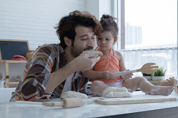 Wall Mural - Caucasian family father and little daughter cooking in the kitchen. Cute girl, face and body full of flour, looking at dad while he eating a piece of bread at home