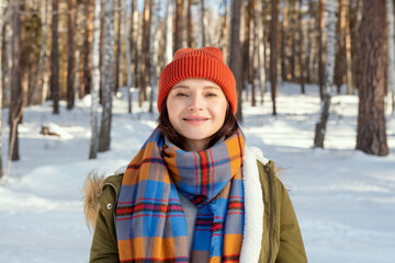 Wall Mural - Young cheerful woman in warm jacket, checkered scarf and beanie looking at you