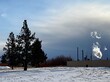 Bright white steam from an industrial plant at the edge of a snow covered field contrasts against a dark and stormy sky.