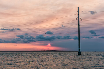Wall Mural - Sunset in Islamorada, Florida Keys with sky power lines pylon in water of Gulf of Mexico or Atlantic Ocean with island horizon by coast shore and sun behind clouds cloudy sky