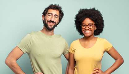 Wall Mural - multiracial couple of friends smiling happily with a hand on hip and confident, positive, proud and friendly attitude