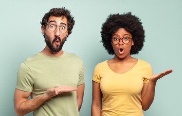 Canvas Print - multiracial couple of friends looking surprised and shocked, with jaw dropped holding an object with an open hand on the side