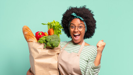 Wall Mural - young pretty afro woman smiling with a happy, confident expression with hand on chin, wondering and looking to the side and holding a vegetables bag