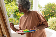 Thoughtful African American Senior Woman Painting On Canvas While Standing On Porch Of The House