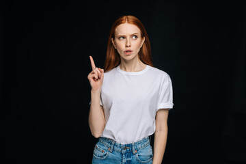 Wall Mural - Red-haired young woman wearing T-shirt and denim having good idea with her finger pointing up isolated black background. Pretty redhead lady model emotionally showing facial expressions.