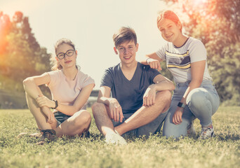 Group of smiling teenage boy and two girls having fun together on summer day, sitting on green grass