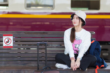 Asian Woman In White Long Sleeve And Hat Sits On Chair With Her Bag At Train Platform In Social Distancing As A New Normal Lifestyle.