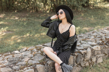 Wall Mural - Comely young woman in sunglasses in beautiful hat in fashionable casual black spring-summer clothes from new stylish youth collection rests on stones on nature on bright sunny day. Trendy girl model.