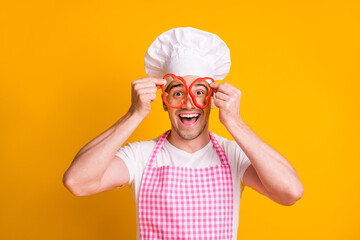 Wall Mural - Photo of young happy excited good mood smiling man cook hold peppers on eyes binoculars isolated on yellow color background