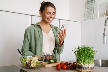 Wall Mural - Happy nice woman using mobile phone while preparing salad