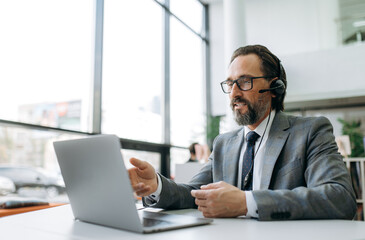 Senior adult business male in headset is sitting at the desk, using laptop, communicate with clients. Caucasian grey-haired manager is consulting people, support service concept