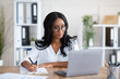 Happy black young business lady working on laptop at office, taking notes during online work meeting or webinar