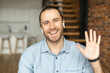 © Vadim Pastuh - Young European man looking at the camera, smiling and waving the hand, positive person with bewitching smile, say hi, with white earphones, greeting family or friends by video, wearing blue shirt