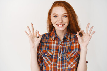 Wall Mural - Young woman with blue eyes and red hair saying yes, showing okay sign and nod in approval, smiling satisfied with good choice, white background