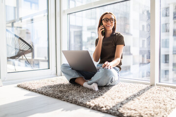 Young woman freelancer working from home, sitting on floor and using laptop, talking with client on phone