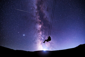 Wall Mural - fantasy night scene. silhouette of a guy swinging on a swing under bright milky way galaxy. beautifu