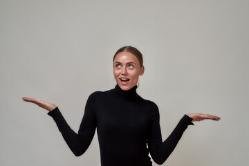 Portrait of surprised beautiful caucasian young woman wearing black turtleneck presenting your very exciting product, standing isolated over gray background