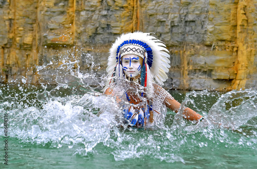 Plakat An Indian girl is seen in a quarry lake. She is dressed
in white and wears a traditional Indian costume with a
feathered headdress on her head, She splashes water
around herself with her hands.