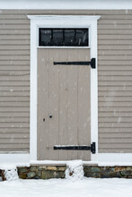 An Old Wooden And Rotten Weathered Beaten Door On The Exterior Of A Tan Colour Clapboard Building With Large Snowflakes Coming Down. There Are Two Large Wrought Iron Hinges On The Left Handed Door.