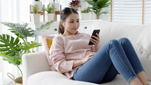 Young Attractive Beautiful Asian Woman Wear Earphone Headphone Listen To Social Media Application Clubhouse Podcast Or Audio Chat App Feeling Happy Comfort Cozy At Home Sitting At Sofa Couch.