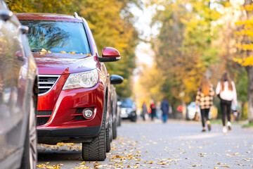 Cars parked in a row on a city street side on bright autumn day with blurred people walking on pedestrian zone.