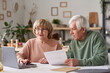 © AnnaStills - Senior couple sitting at the table and using laptop to pay for bills online in the kitchen