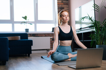 Relaxed redhead young woman with closed eyes meditating at home sitting in lotus position on yoga mat near laptop computer. Peaceful red-haired lady holding hands on knee in Om position.