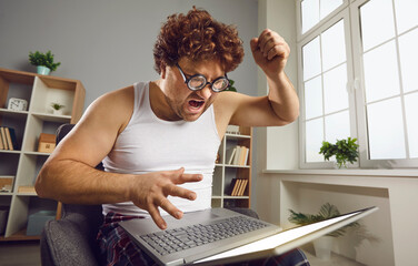 Funny angry curly guy having problem with his computer at home. Furious crazy nerdy young man experiencing software failure, shouting and holding fist over laptop threatening to break it