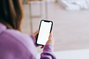 Woman holding a smartphone with a white screen mock up, resting on the armchair in living room at home.