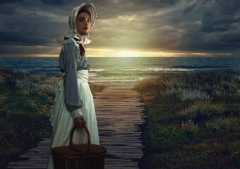 Wall Mural - Victorian girl with hood and wicker basket in white dress and blue striped blouse on the coast at sunset.