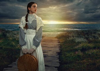 Wall Mural - Victorian girl with a hood and a wicker basket in her hands dressed in a white dress and blue striped blouse stands on the seashore at sunset.