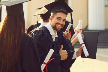 Portrait of a graduate student with the diploma and in the black academic gown raises his thumbs up at graduation at university. Student looks at the camera and smiles sincerely.