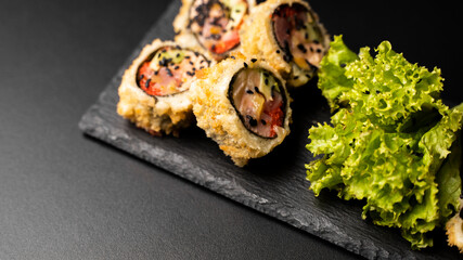 Wall Mural - Custom sushi roll in tempura with nori, fresh salmon, tuna, avocado, masago caviar, drizzled with pineapple sauce with salad pouring as decoration on a black plate on a black table and background.