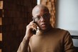 © Serhii - African businessman talking on phone sitting at cafe table, busy entrepreneur working distantly in coffee house with laptop papers speaking on mobile, black man making call having lunch in cafeteria
