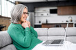 © Vadim Pastuh - Smiling and excited senior woman sitting on the couch in cozy living room at home, using computer laptop for spending leisure time, lovely grandmother received photo of grandchildren