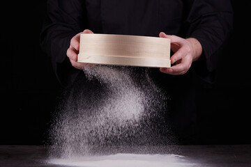 chef a man in a black uniform holds a round wooden sieve in his hands and sifts white wheat flour on a black background