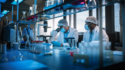 Modern Medical Research Laboratory: Two Scientists Wearing Face Masks use Microscope, Analyse Sample in Petri Dish, Discuss Innovative Technology. Advanced Scientific Lab for Medicine, Biotechnology