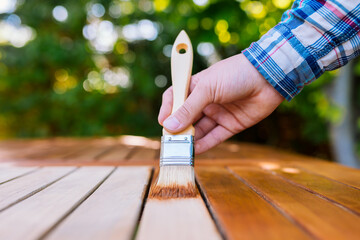 a hand holding a brush applying varnish paint on a wooden garden table - painting and caring for wood with oil