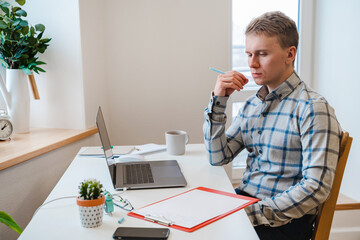 Canvas Print - a young man in a shirt in a bright office works at a laptop and makes notes in a notebook, the menta