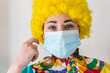 © AliciaFdez - Linda mujer joven sonriendo mientras está vestida con un disfraz de payaso con una mascarilla quirúrgica desechable y una peluca de payaso amarilla. Protección contra el virus sars-cov-2