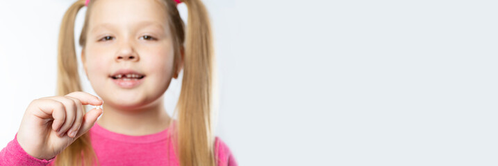 Little caucasian girl with long blond hair in pink clothes holds a pulled out milk tooth in her hands on a light background