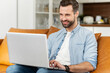 © Vadim Pastuh - Smiling young bearded hipster man sitting on the couch, flirting, typing a message on the laptop, video-calling with friends or girlfriend online, chatting in social media network, using dating app