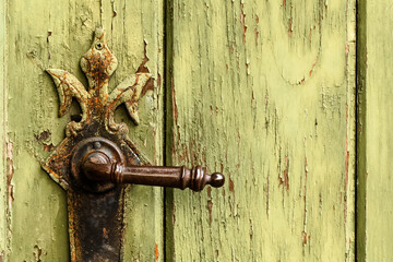 Old rusty door handle on green door. Detail of rustic old vintage and rusty grungy iron door handle installed on an ornamented old heavy and massive medieval green wooden door with peeling paint.