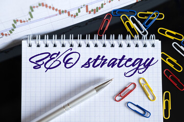 On the desktop are a forex chart, paper clips, a pen and a notebook in which it is written - SEO strategy