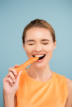 Cheerful Young Woman With Natural Makeup Biting Whole Carrot Isolated On Blue