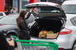 © Aleksandr - Young stylish woman in protective mask with shopping cart full of fresh food during quarantine, packing products into the car on the outdoor parking. Woman close car trunk on parking
